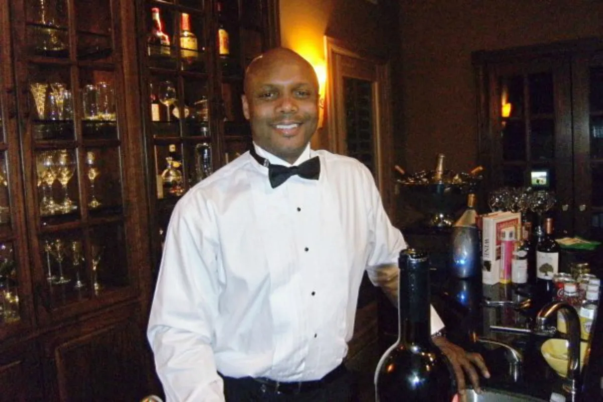 Eric Chapman in a tuxedo shirt and bow tie beside a wine bottle at a dark wood bar