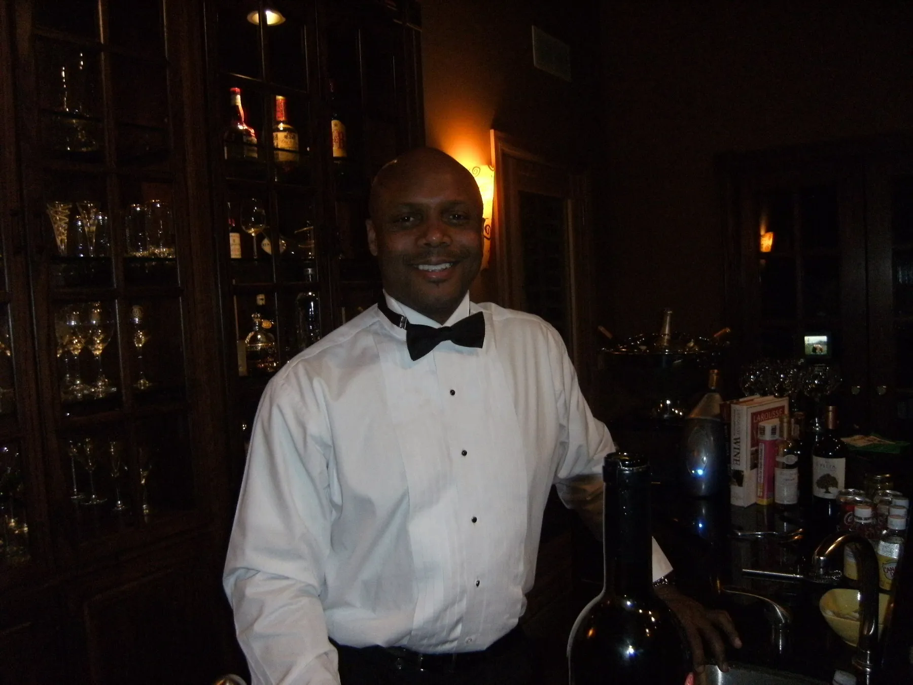 Eric Chapman in a tuxedo shirt and bow tie beside a wine bottle at a dark wood bar