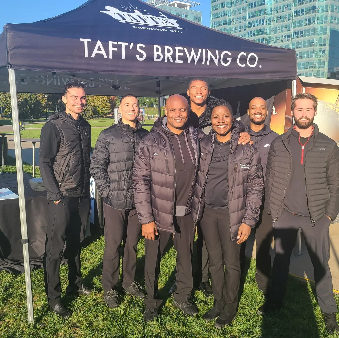 Seven First Call Bartending staff in matching black jackets under a brewery tent at a University of Cincinnati homecoming party